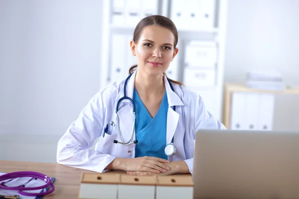 Schöne junge lächelnde Ärztin sitzt am Schreibtisch und schreibt. — Stockfoto