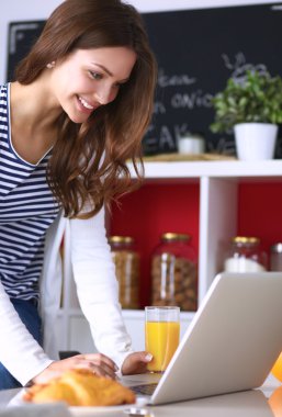 Dizüstü bilgisayar kullanarak ve mutfakta oturmuş çekici genç kadın