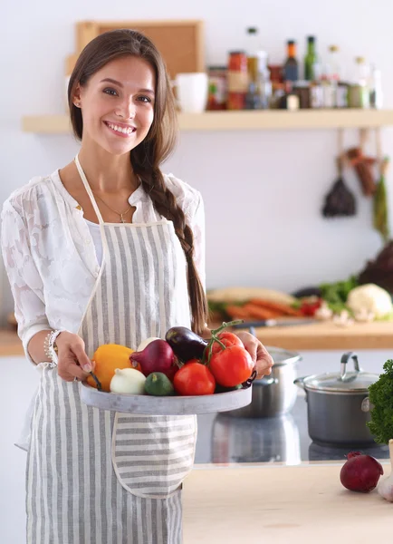 Mutfakta elinde sebzelerle gülümseyen genç bir kadın.