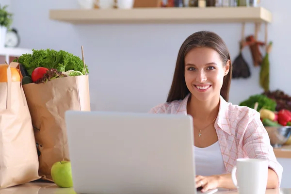 Lächelnde junge Frau mit Kaffeetasse und Laptop in der heimischen Küche — Stockfoto