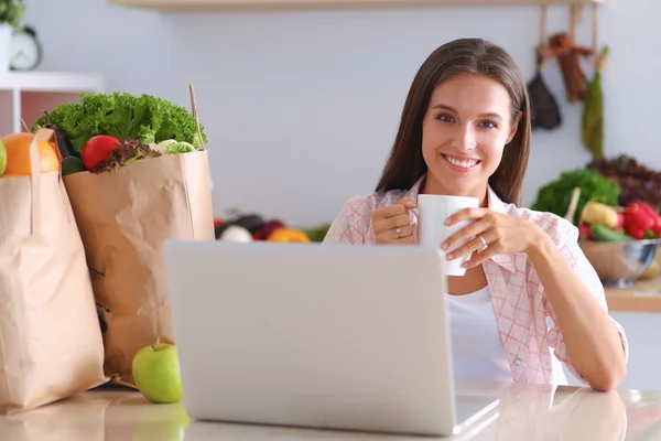 コーヒー カップと自宅の台所でラップトップを持つ若い女性の笑みを浮かべてください。 — ストック写真