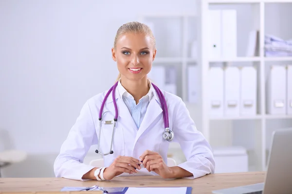 Mooie jonge glimlachende vrouwelijke arts zitten aan het bureau en schrijven. — Stockfoto