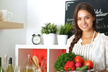 Mutfakta elinde sebzelerle gülümseyen genç bir kadın.
