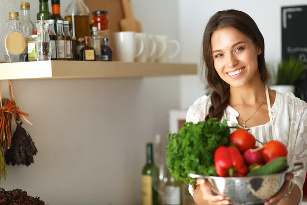 Mujer joven sonriente sosteniendo verduras de pie en la cocina —  Fotos de Stock