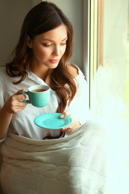 Evde oturan genç bir kadın oturma odasında oturmuş kitap okuyor ve çay ya da kahve içiyor.