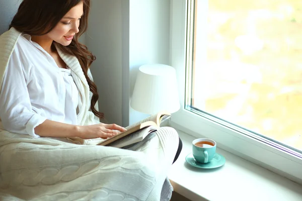 Young woman at home sitting near window relaxing in her living room reading book and drinking coffee or tea