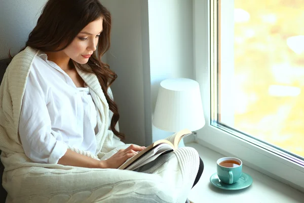 Jonge vrouw thuis zitten in de buurt van raam ontspannen in haar woonkamer lezen boek en het drinken van koffie of thee — Stockfoto