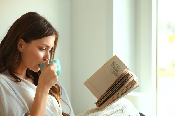 Mujer joven en casa sentada cerca de la ventana relajándose en su sala de estar leyendo libro y tomando café o té —  Fotos de Stock