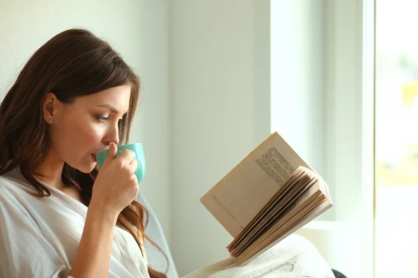 Young woman at home sitting near window relaxing in her living room reading book and drinking coffee or tea