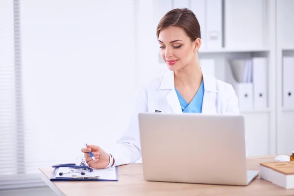 Mooie jonge glimlachende vrouwelijke arts zitten aan het bureau en schrijven. — Stockfoto