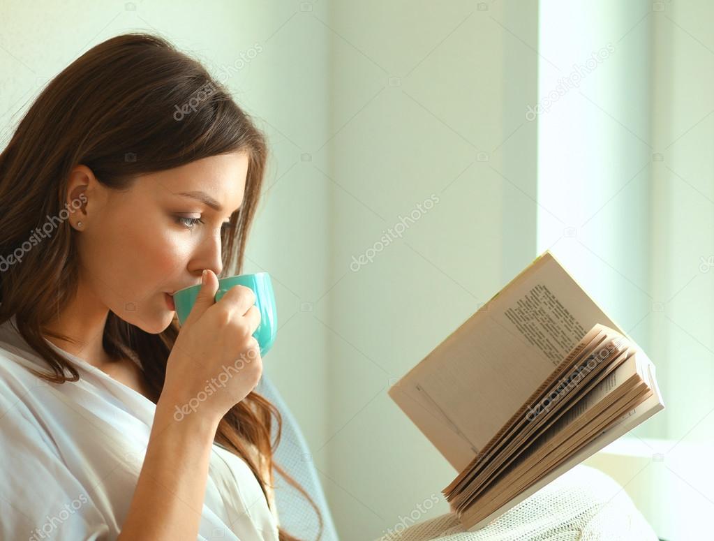 Evde oturan genç bir kadın oturma odasında oturmuş kitap okuyor ve çay