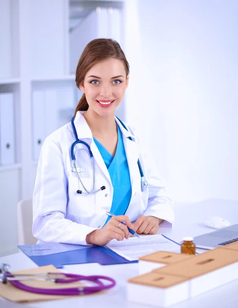 Mooie jonge glimlachende vrouwelijke arts zitten aan het bureau en schrijven. — Stockfoto