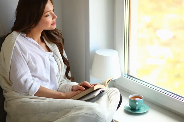 Jonge vrouw thuis zitten in de buurt van raam ontspannen in haar woonkamer lezen boek en het drinken van koffie of thee — Stockfoto