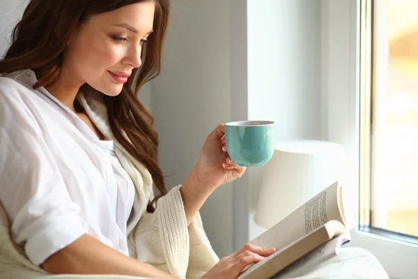 Tânăra de acasă stând lângă fereastră relaxându-se în sufrageria ei citind cartea și bând cafea sau ceai — Fotografie, imagine de stoc