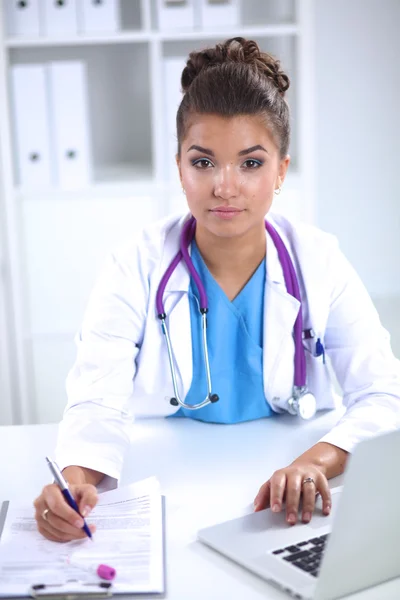 Hermosa joven sonriente doctora sentada en el escritorio y escribiendo. —  Fotos de Stock