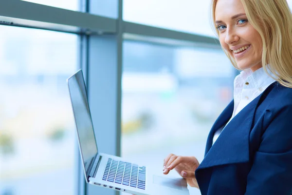 Donna d'affari in piedi contro la finestra dell'ufficio che tiene il computer portatile — Foto Stock