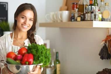 Mutfakta elinde sebzelerle gülümseyen genç bir kadın.