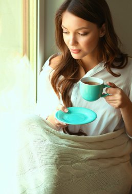 Evde oturan genç bir kadın oturma odasında oturmuş kitap okuyor ve çay ya da kahve içiyor.