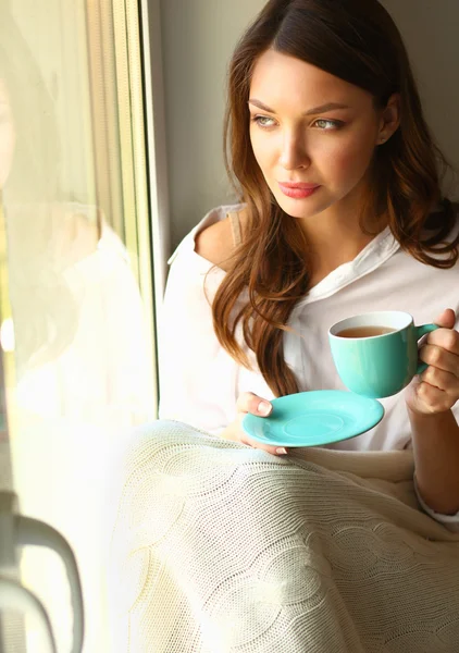 Jonge vrouw thuis zitten in de buurt van raam ontspannen in haar woonkamer lezen boek en het drinken van koffie of thee — Stockfoto