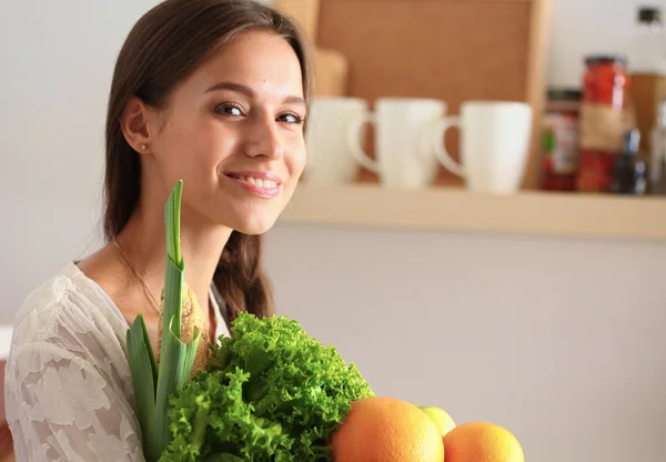 Leende ung kvinna som håller grönsaker stående i köket — Stockfoto
