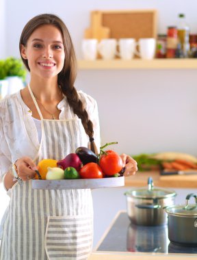 Mutfakta elinde sebzelerle gülümseyen genç bir kadın.