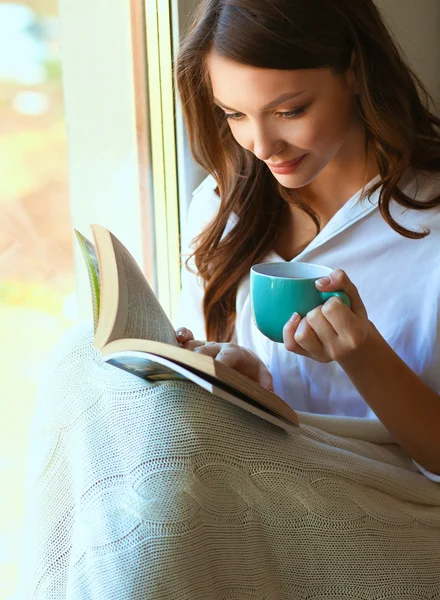 Jonge vrouw thuis zitten in de buurt van raam ontspannen in haar woonkamer lezen boek en het drinken van koffie of thee — Stockfoto