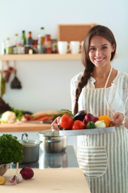 Mutfakta elinde sebzelerle gülümseyen genç bir kadın.