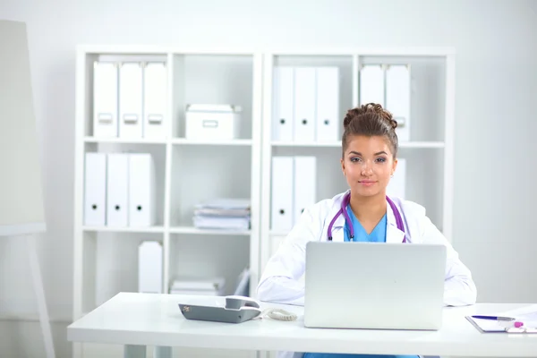 Mooie jonge glimlachende vrouwelijke arts zitten aan het bureau en schrijven. — Stockfoto