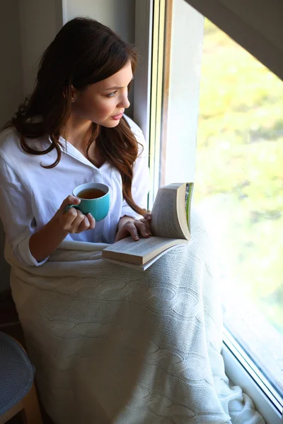 Jonge vrouw thuis zitten in de buurt van raam ontspannen in haar woonkamer lezen boek en het drinken van koffie of thee — Stockfoto