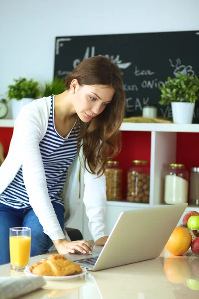 Mujer joven atractiva usando el ordenador portátil y sentado en la cocina —  Fotos de Stock