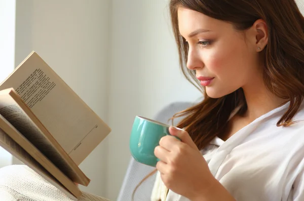 Jovem mulher em casa sentado perto da janela relaxante em sua sala de estar leitura livro e beber café ou chá — Fotografia de Stock