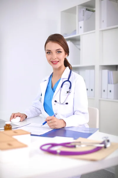 Hermosa joven sonriente doctora sentada en el escritorio y escribiendo. — Foto de Stock