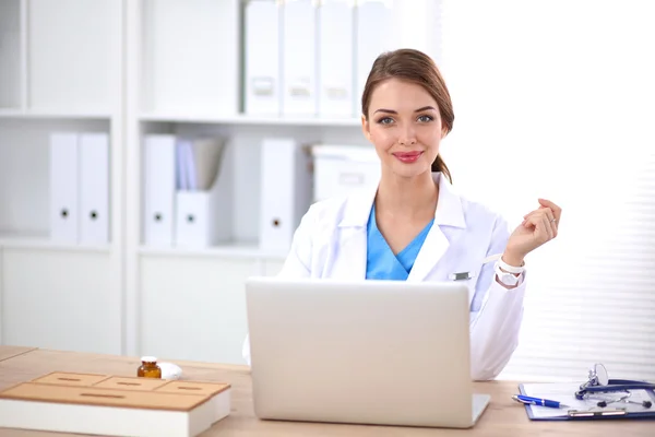 Hermosa joven sonriente doctora sentada en el escritorio y escribiendo. — Foto de Stock