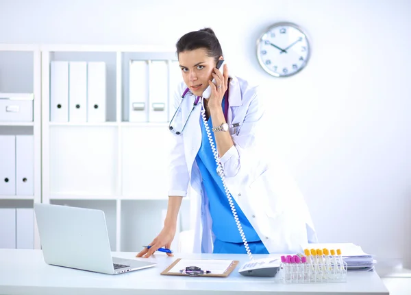 Young woman doctor in white coat at computer using phone — Stock Photo, Image