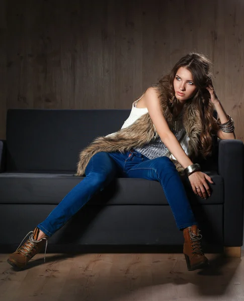 Retrato de una mujer elegante sentada en un sofá negro con pantalones vaqueros azules y chaleco de piel —  Fotos de Stock