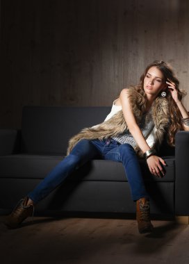 Mavi jeans giydiği siyah koltukta oturan zarif kadın portresi ve kürk yelek