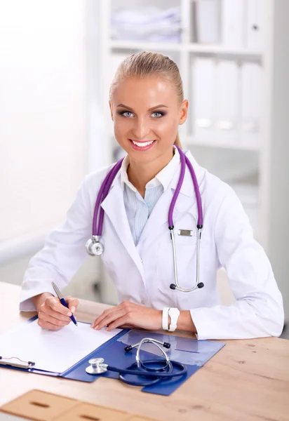 Beautiful young smiling female doctor sitting at the desk and writing. — Stock Photo, Image