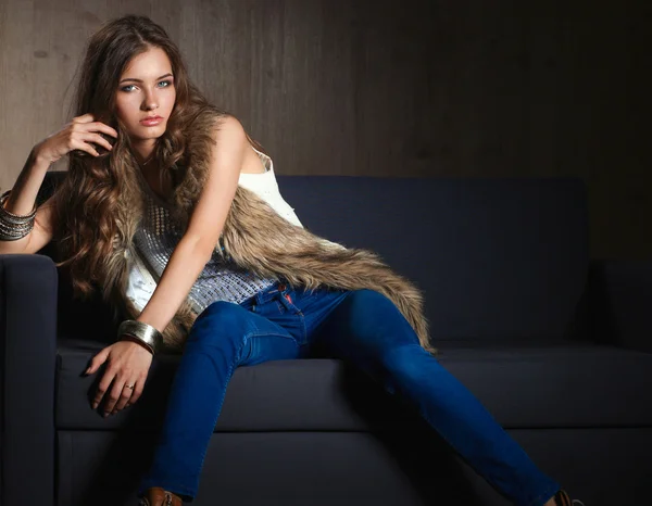 Retrato de mulher elegante sentado no sofá preto vestindo uma calça azul e colete de pele — Fotografia de Stock