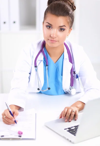 Schöne junge lächelnde Ärztin sitzt am Schreibtisch und schreibt. — Stockfoto