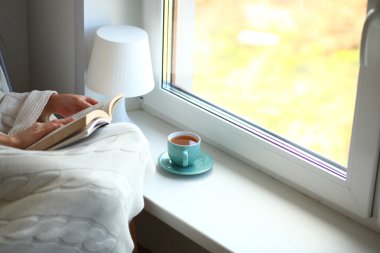 Evde oturan genç bir kadın oturma odasında oturmuş kitap okuyor ve çay ya da kahve içiyor.