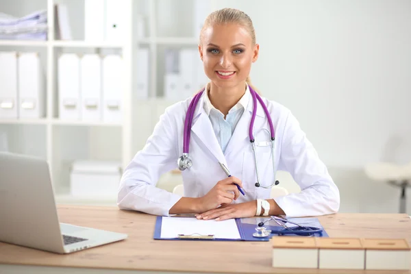 Bonito jovem sorridente médico feminino sentado na mesa e escrevendo. — Fotografia de Stock