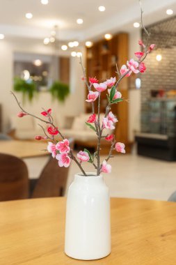 Yapay Japon sakura bitkisi büyük bir salonda, ahşap masanın üzerinde beyaz bir vazoda.