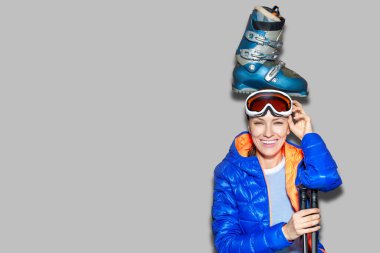 Kışlık giysiler, gözlük takan, elinde kayak sopası ve evde kayak botu olan bir kız.