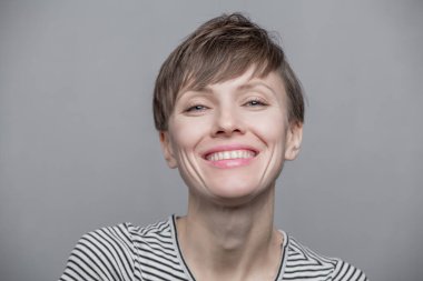 Kısa saçlı, neşeli genç bir kadın açık gri arka planda doğal makyaj yapıyor. Güzel esmer bir kadının kapalı bir portresi.