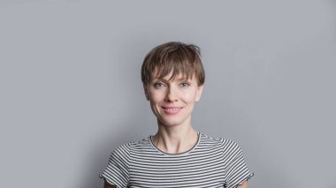 Kısa saçlı, neşeli genç bir kadın açık gri arka planda doğal makyaj yapıyor. Güzel esmer bir kadının kapalı bir portresi.