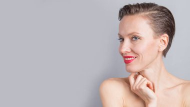 Kısa saçlı, neşeli genç bir kadın açık gri arka planda doğal makyaj yapıyor. Güzel esmer bir kadının kapalı bir portresi.