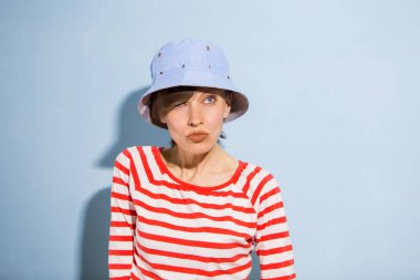 Kısa saçlı mutlu bir genç kadın, açık mavi arka planda doğal makyaj. Güzel esmer, mavi şapkalı ve parlak çizgili elbiseli bir kadının kapalı bir portresi.