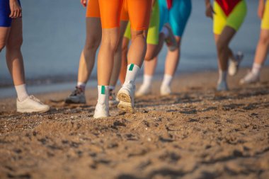 İnce vücutlu, sportif aktif kadınlardan oluşan bir grup. Sahilde spor yapıyorlar. Parlak mayo giyiyorlar.