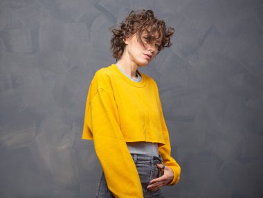 Kısa kıvırcık saçlı, parlak sarı süveter giyen, gri arka planda doğal makyajı olan mutlu, genç bir kadın.