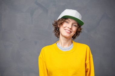 Sonbahar havası! Kısa esmer, kıvırcık saçlı, sarı kazaklı ve gri duvarlı arka planda şapkası olan mutlu bir kadının portresi.